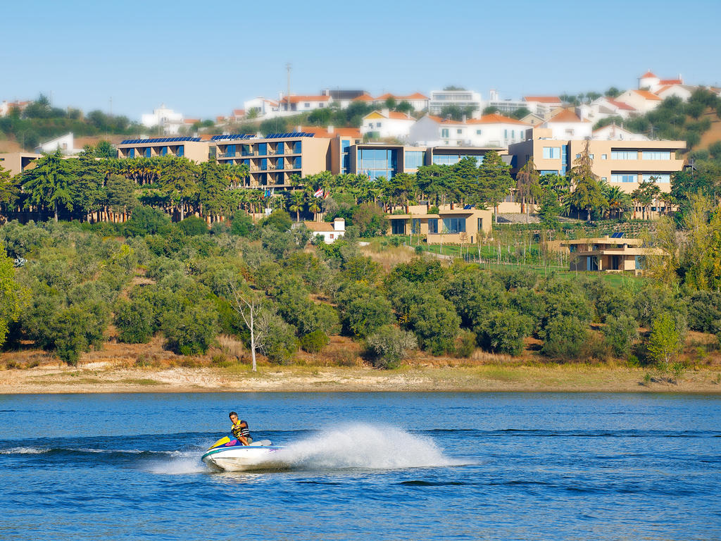 Ap Lago Montargil - Conference & Spa エクステリア 写真 The photo depicts a person riding a jet ski on a body of water. In the background, there are modern buildings and lush greenery, suggesting a scenic landscape, likely near a resort or upscale residential area. The clear blue sky and calm water enhanc