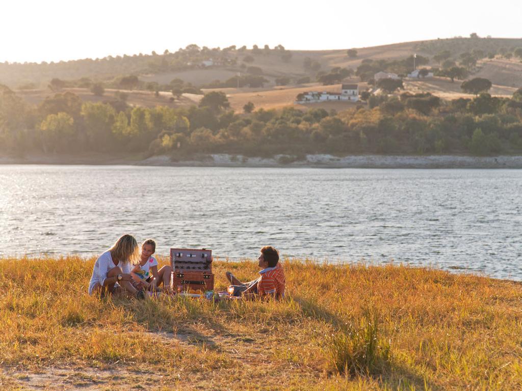 Ap Lago Montargil - Conference & Spa エクステリア 写真 The photo depicts a serene lakeside scene during sunset. Three people are gathered on a grassy area near the water, enjoying a picnic. They are seated around a picnic basket, with the shimmering lake in the background and hills in the distance. The w