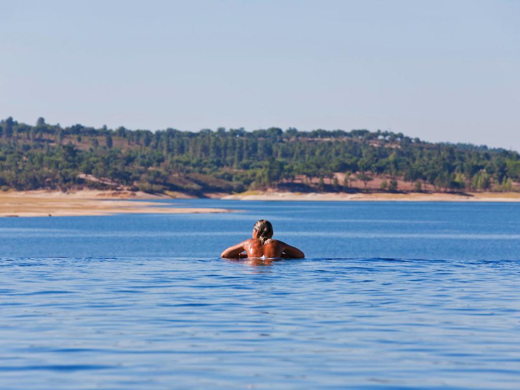 Ap Lago Montargil - Conference & Spa エクステリア 写真 The photo shows a person swimming in a calm body of water, likely a lake or a river. The swimmer is seen from behind and appears to be enjoying the water on a sunny day. In the background, there's a scenic landscape featuring green trees and a sandy 