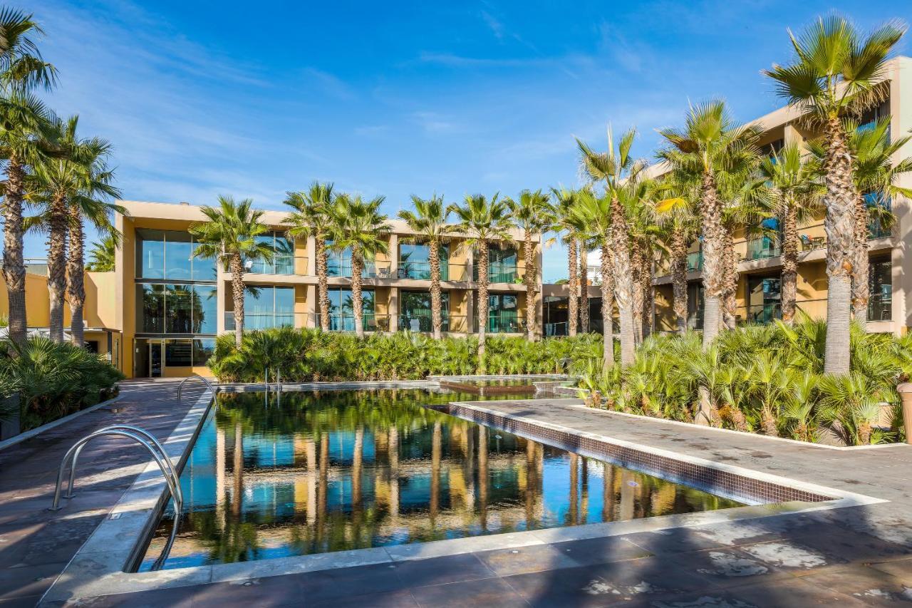 Ap Lago Montargil - Conference & Spa エクステリア 写真 The photo features a modern building surrounded by lush greenery and palm trees. In the foreground, there is a serene swimming pool with a reflective surface that captures the surrounding environment. The architecture of the building appears to have 