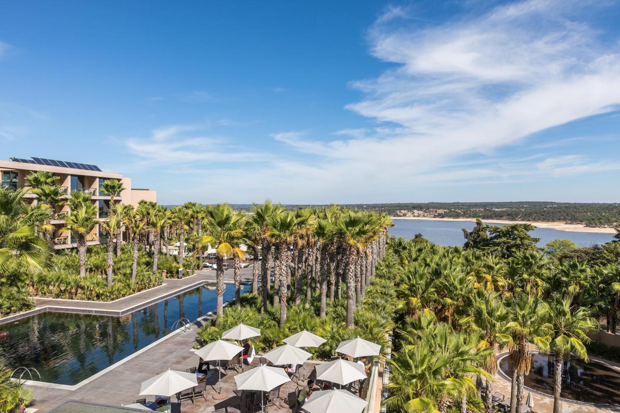 Ap Lago Montargil - Conference & Spa エクステリア 写真 The photo shows a scenic view of a resort or hotel area. In the foreground, there is a pool surrounded by palm trees and lounge chairs with umbrellas, suggesting a relaxing atmosphere. Beyond the pool, there is a body of water, likely a river or coas
