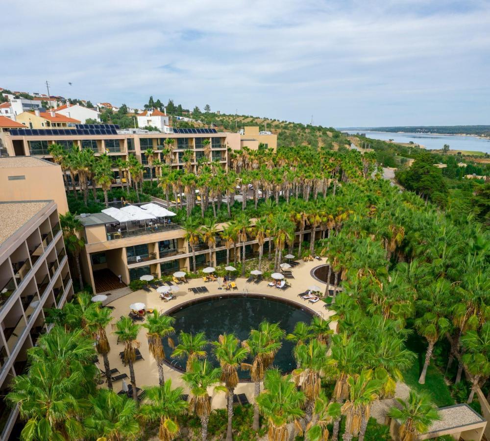 Ap Lago Montargil - Conference & Spa エクステリア 写真 The photo shows a modern hotel complex set amidst lush greenery, featuring numerous palm trees. In the foreground, there is a circular swimming pool surrounded by lounge chairs. The hotel itself has multiple levels with balconies overlooking the pool