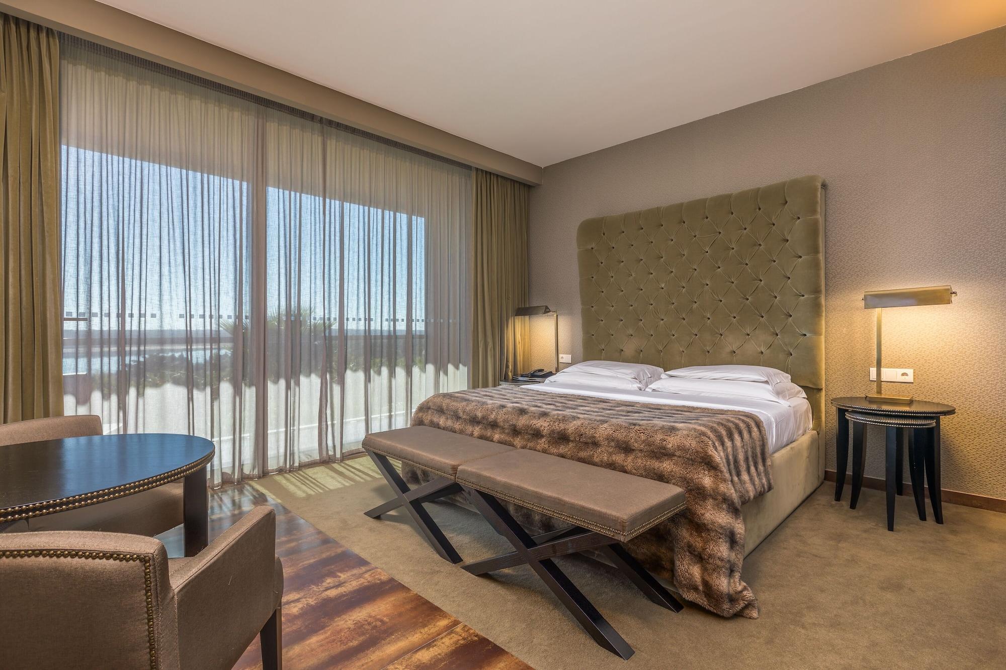 Ap Lago Montargil - Conference & Spa エクステリア 写真 The photo shows a modern hotel room featuring a large bed with a tufted headboard and a cozy blanket. There are curtains draped over large windows that allow natural light to flow in, providing a view of the outside. A small seating area includes two
