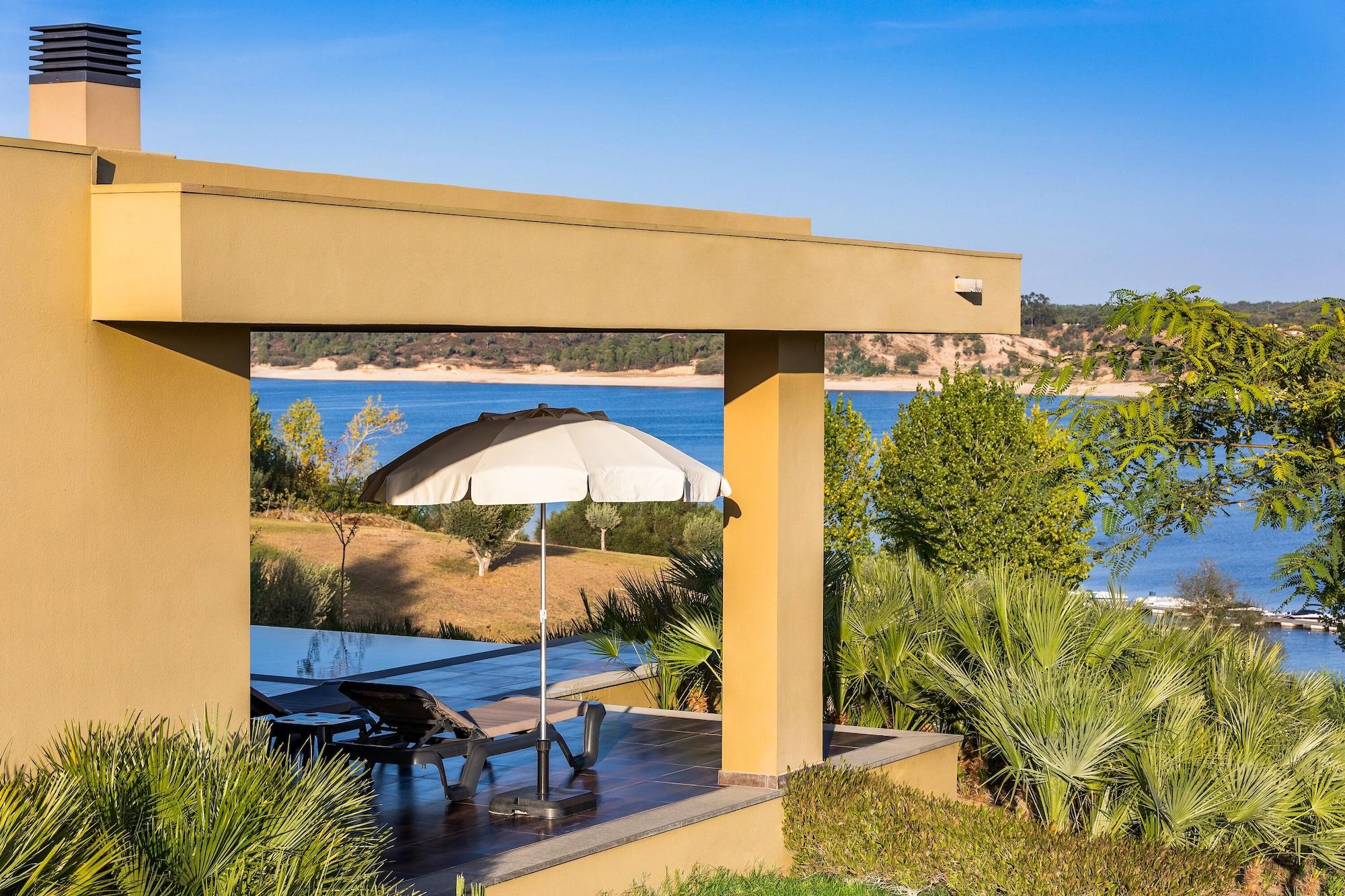 Ap Lago Montargil - Conference & Spa エクステリア 写真 The photo shows a peaceful outdoor area with a modern architectural structure featuring an overhang. There is a lounge chair positioned beneath a large umbrella, creating a shaded spot to relax. In the background, there is a picturesque view of a bod