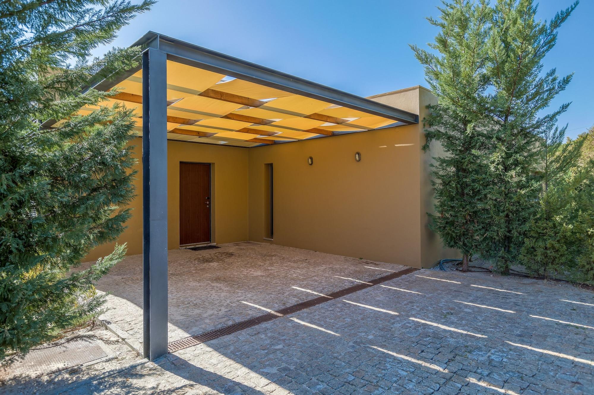 Ap Lago Montargil - Conference & Spa エクステリア 写真 The photo shows an exterior view of a modern structure with a carport. The carport has a sleek, metallic frame and a yellowish canopy above it. In front of the structure, there is a paved area made of stone or tiles. The main entrance features a wood