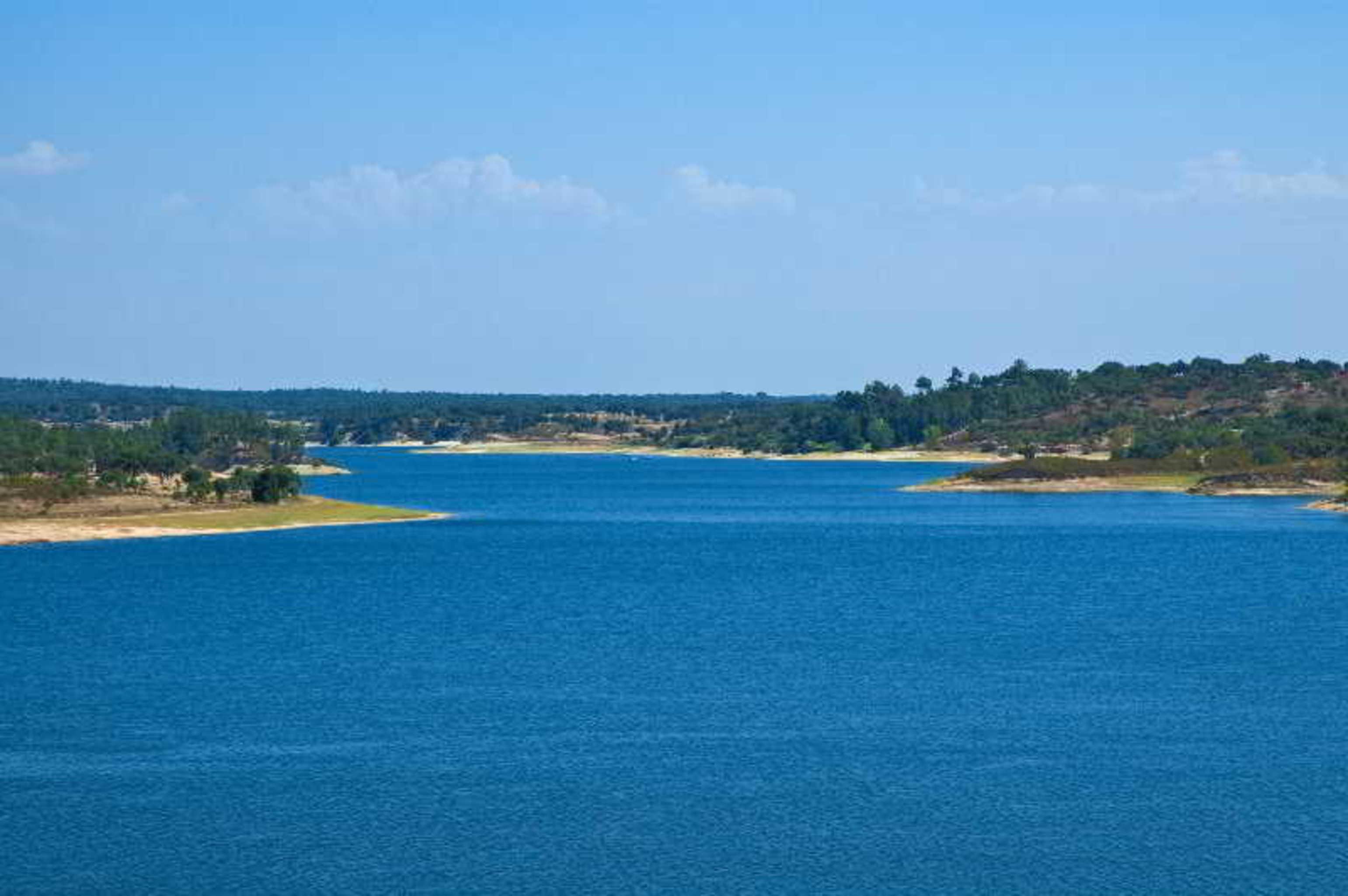 Ap Lago Montargil - Conference & Spa エクステリア 写真 The photo depicts a serene landscape featuring a large body of water, likely a lake or river. The water appears calm and is reflecting a clear blue sky with some clouds. Surrounding the water, there are patches of green land and trees, indicating a n