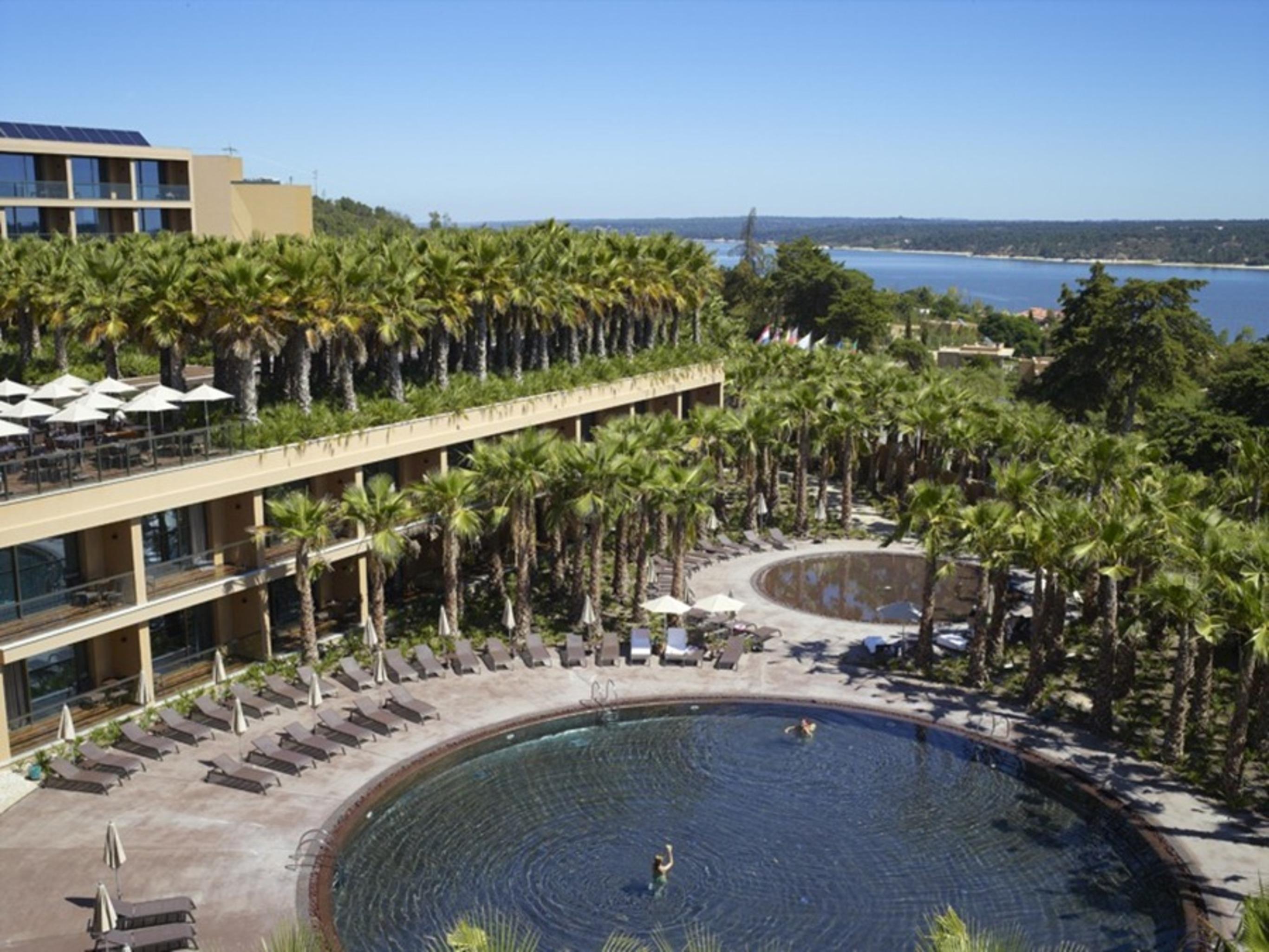 Ap Lago Montargil - Conference & Spa エクステリア 写真 The photo shows a scenic view of a resort area. In the foreground, there are circular pools surrounded by lounge chairs, with a few people enjoying the water. Palm trees line the edges, creating a tropical atmosphere. In the background, there is a mu