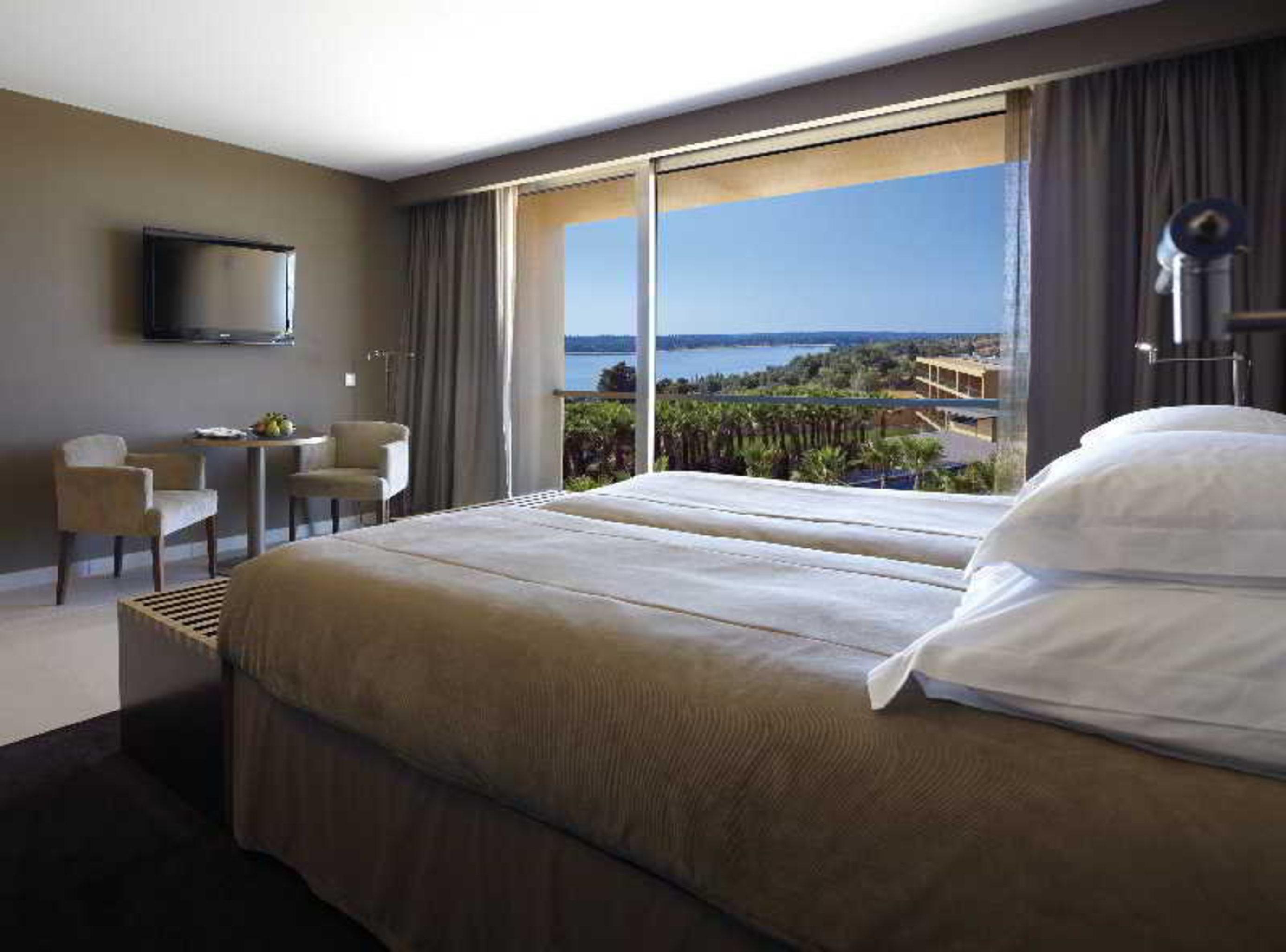 Ap Lago Montargil - Conference & Spa エクステリア 写真 The photo shows a modern hotel room with a large window offering a view of the sea and mountains. The room features a comfortable bed with white pillows and brown bedding. There is a small sitting area with two chairs and a table, as well as a wall-m