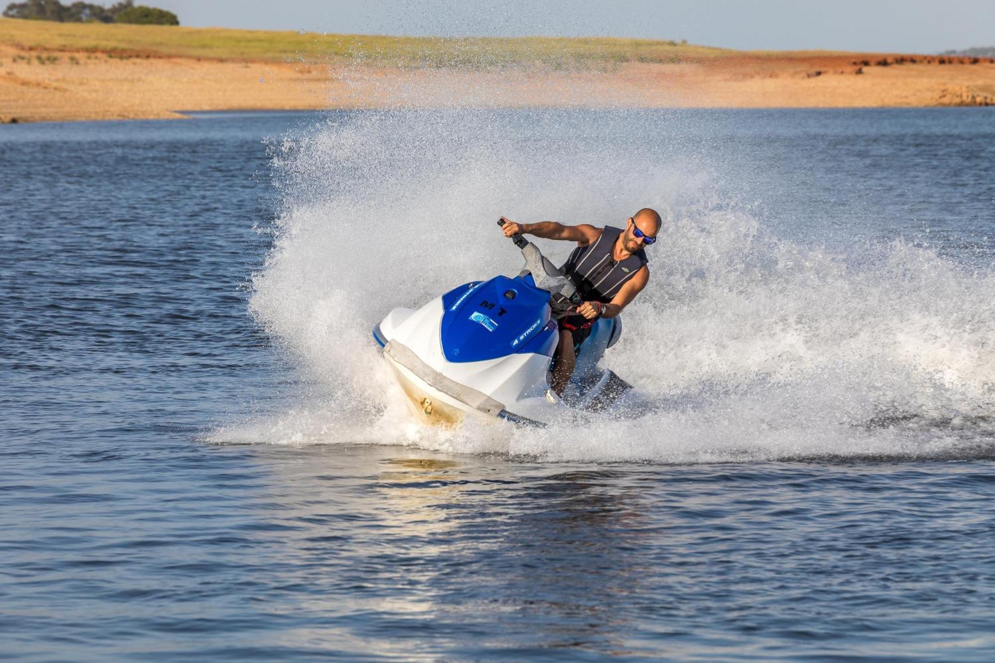 Ap Lago Montargil - Conference & Spa エクステリア 写真 The photo shows a person riding a jet ski over a body of water. The rider is wearing sunglasses and a life vest, leaning forward as the jet ski accelerates, creating a spray of water around them. The background features a natural landscape, likely a 