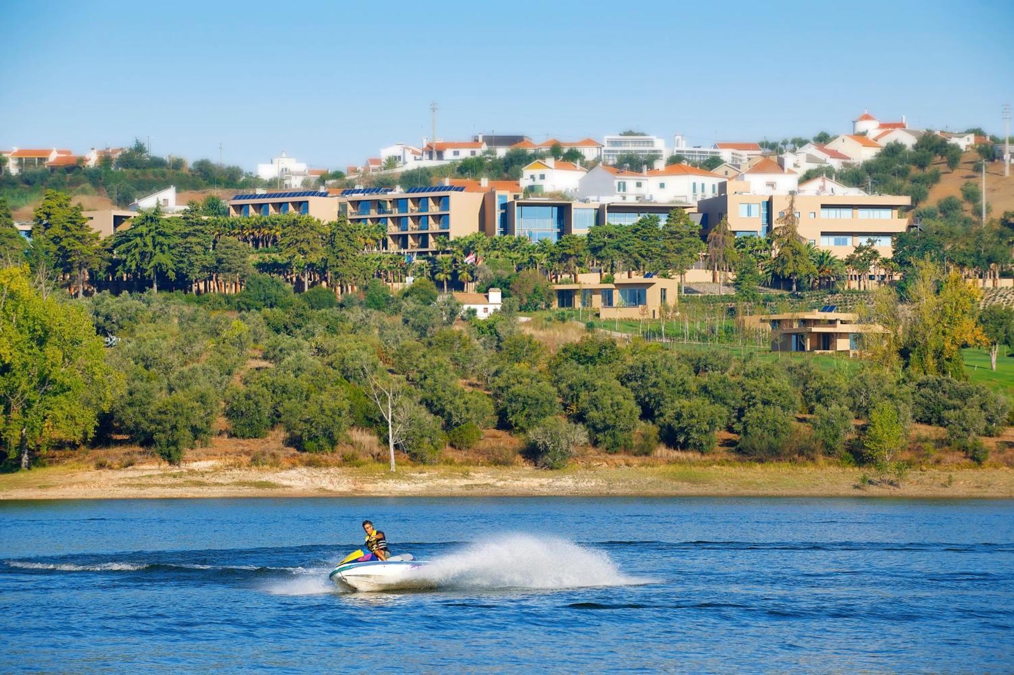 Ap Lago Montargil - Conference & Spa エクステリア 写真 The photo shows a person riding a jet ski on a body of water, creating a spray as they speed across the surface. In the background, there are modern buildings and a hillside with trees and greenery. The sky is clear and blue, suggesting a bright and 