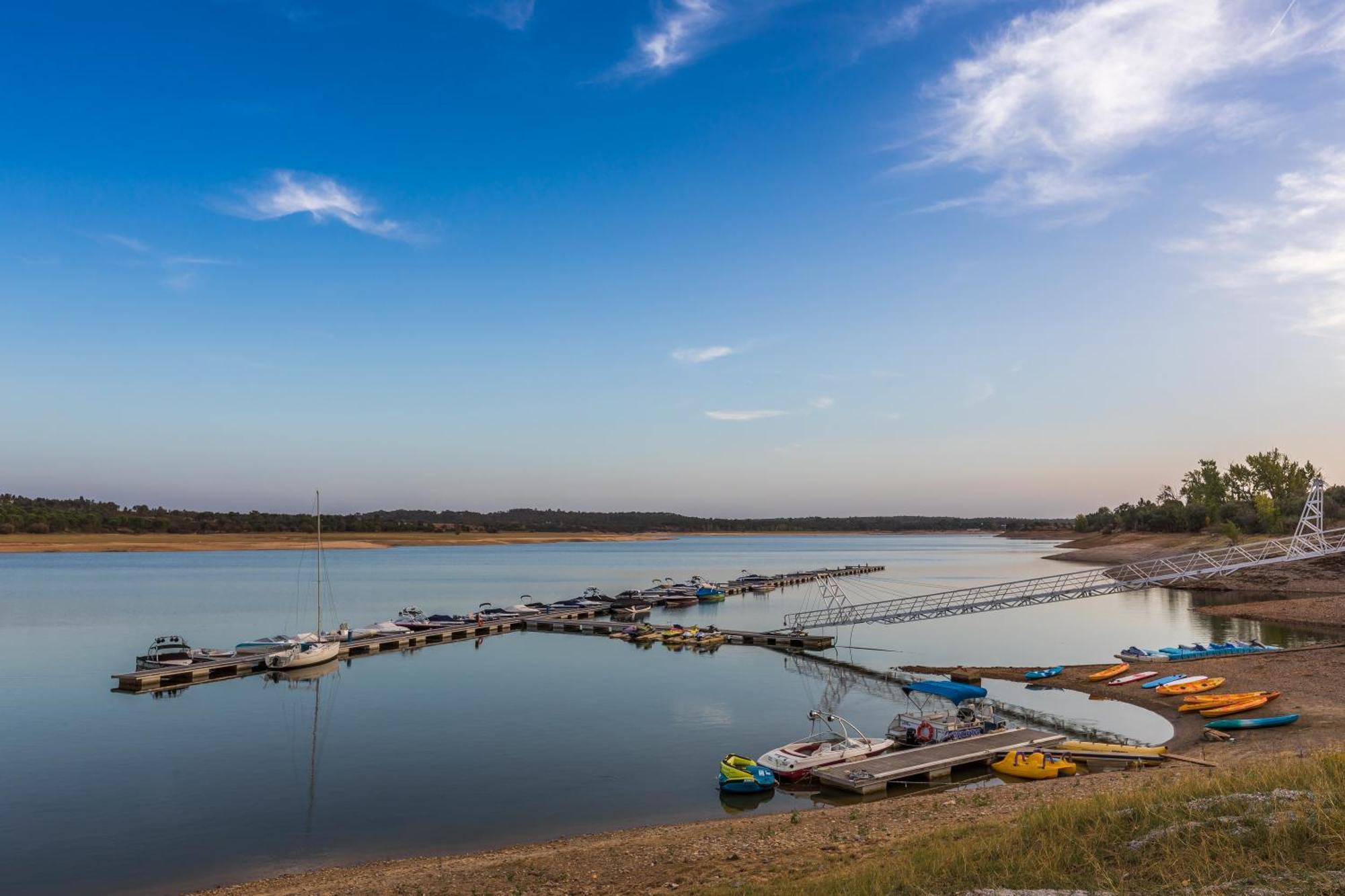 Ap Lago Montargil - Conference & Spa エクステリア 写真 The photo shows a serene lakeside scene. In the foreground, there are several boats moored at a dock, with some colorful kayaks and canoes lined up along the shore. The water is calm and reflects the sky, which features a few wispy clouds. In the bac