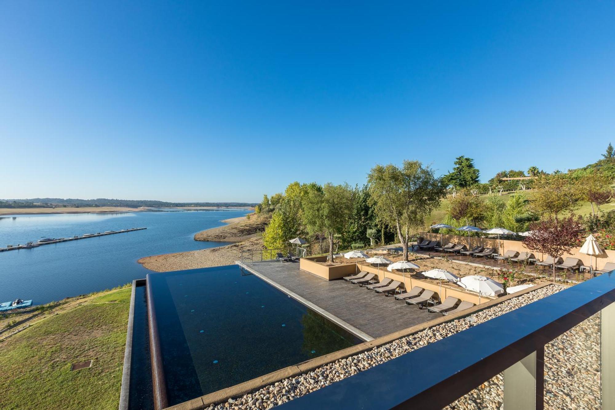 Ap Lago Montargil - Conference & Spa エクステリア 写真 The photograph depicts a scenic view of a tranquil water body, likely a lake or river, bordered by lush greenery. In the foreground, there is an infinity pool that blends seamlessly with the water beyond. Comfortable lounge chairs are arranged on a w