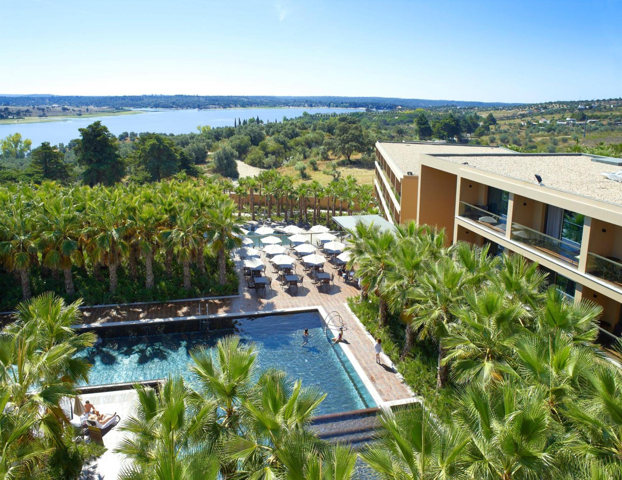 Ap Lago Montargil - Conference & Spa エクステリア 写真 The photo shows a scenic view of a modern hotel or resort. In the foreground, there is a swimming pool surrounded by palm trees, with lounge chairs positioned beside it. Sun umbrellas provide shade over the chairs. The background features a lush land